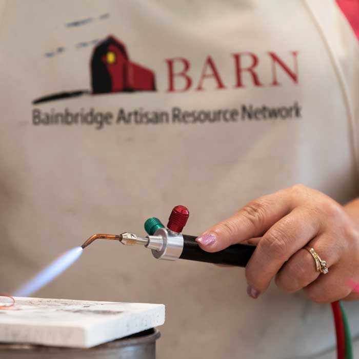 Person wearing a BARN apron and holding an active jeweler's torch